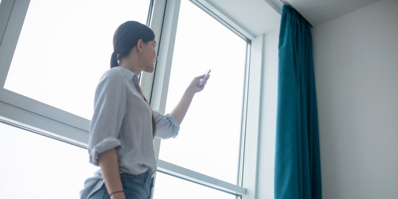 A woman uses a remote control to open smart curtains in a modern, well-lit room.